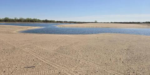 Wide view of a sandy shoreline and shallow blue water at the Newark West Sandpit in Nebraska, with a tree line in the distance under a clear sky.
