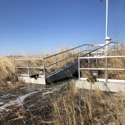Water flowing beneath a metal access platform and hydraulic control structure beside tall grasses.